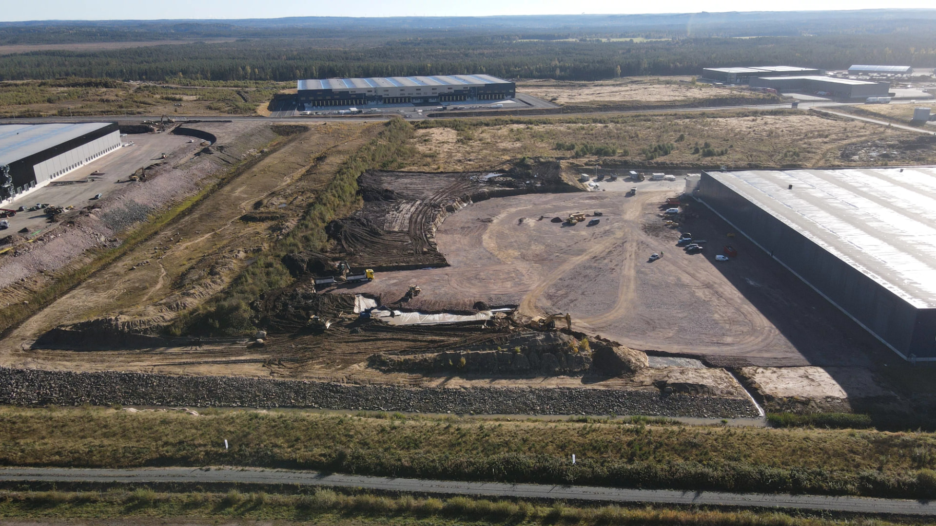 Flygbild över markarbeten vid ett logistikområde med lagerbyggnader och skog i bakgrunden
